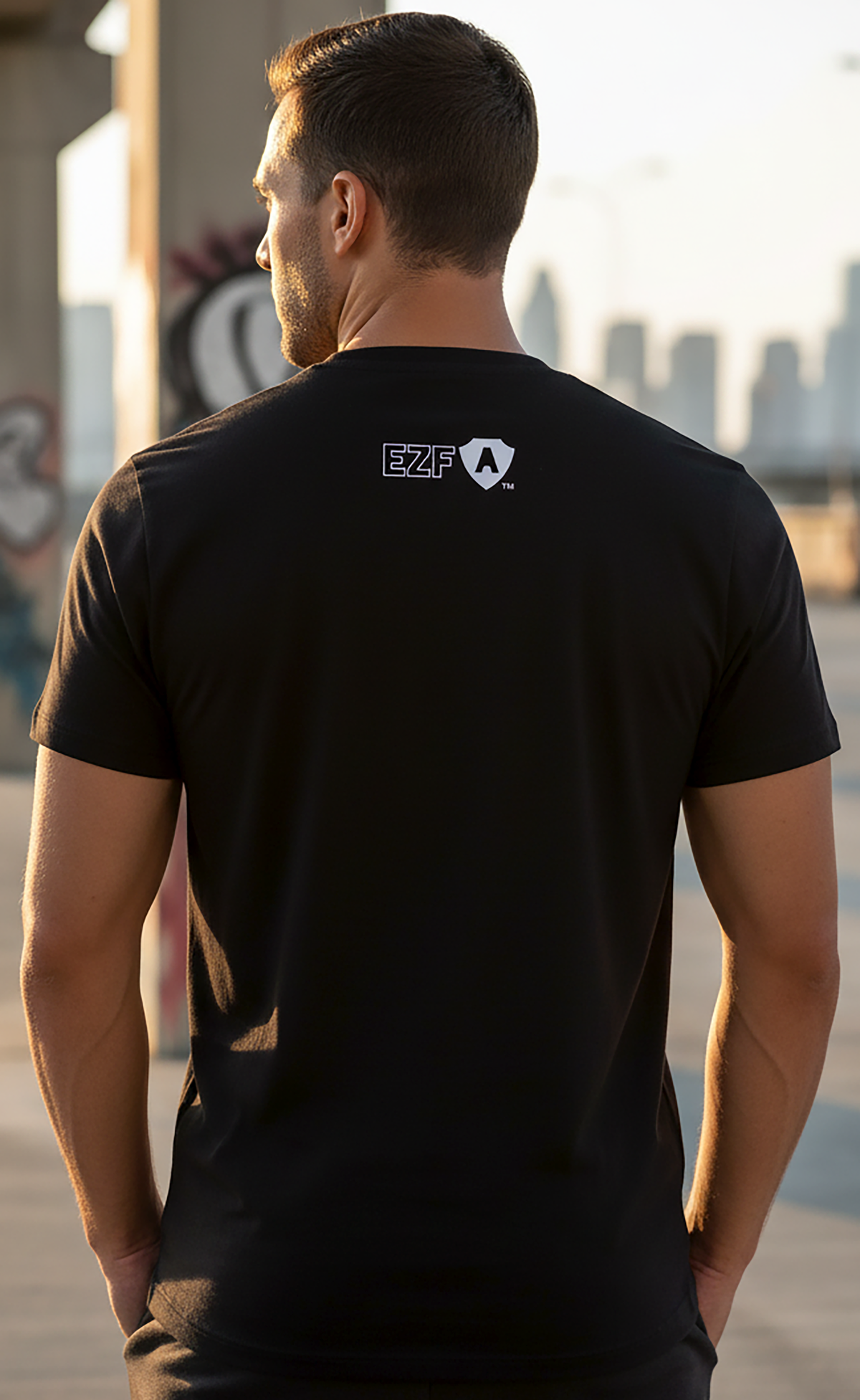 A man viewed from behind wearing a black short-sleeve t-shirt with a white EZF Armor logo printed on the upper back, set against a blurred outdoor urban background.