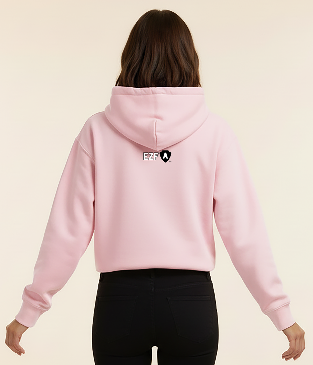 A woman viewed from the back wearing a light pink cropped hoodie with a small black and white graphic logo on the center of the back, paired with black pants, against a solid cream background.