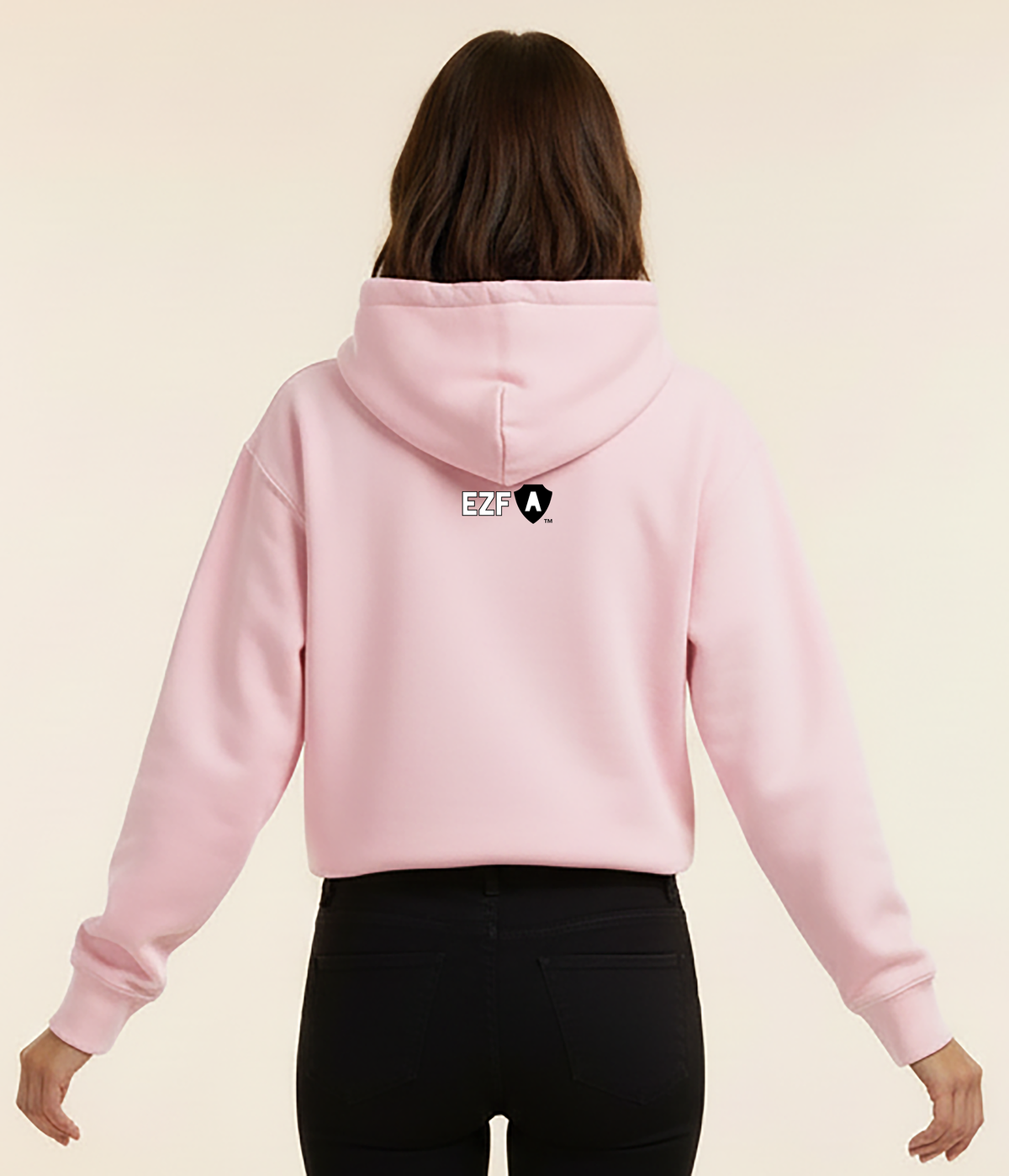 A woman viewed from the back wearing a light pink cropped hoodie with a small black and white graphic logo on the center of the back, paired with black pants, against a solid cream background.